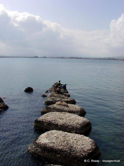 Fishermen on the pier in black volcanic stones, Syracuse, Sicily - Italy